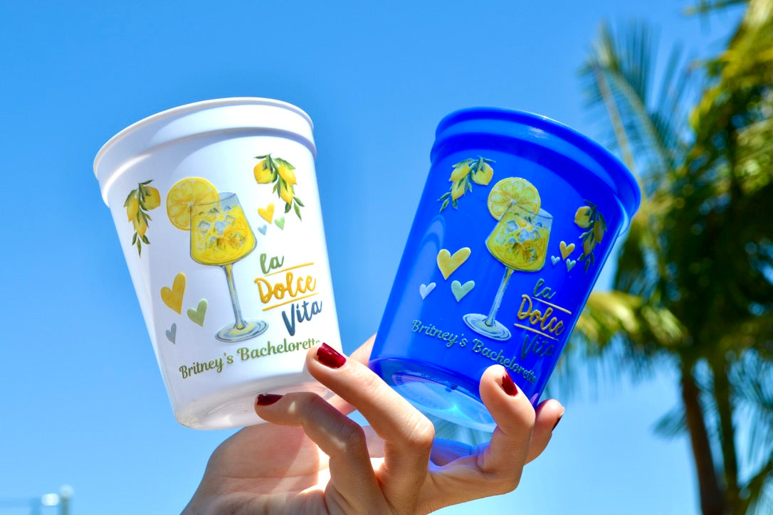 Two branded cups, one white and one blue, held against a clear blue sky with palm trees.