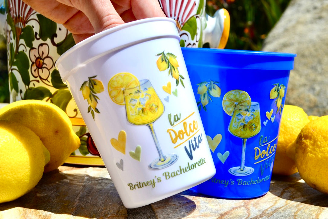 Two plastic cups with lemon-themed designs and text, surrounded by lemons on a stone surface.