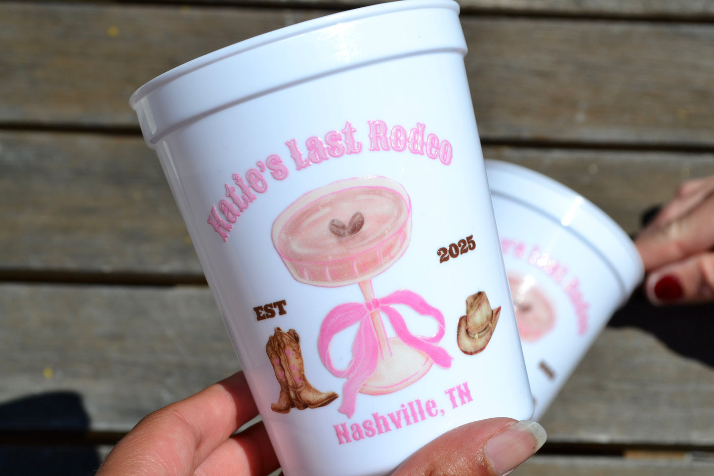White plastic cups with pink decorations and text, held by a person on a wooden surface.