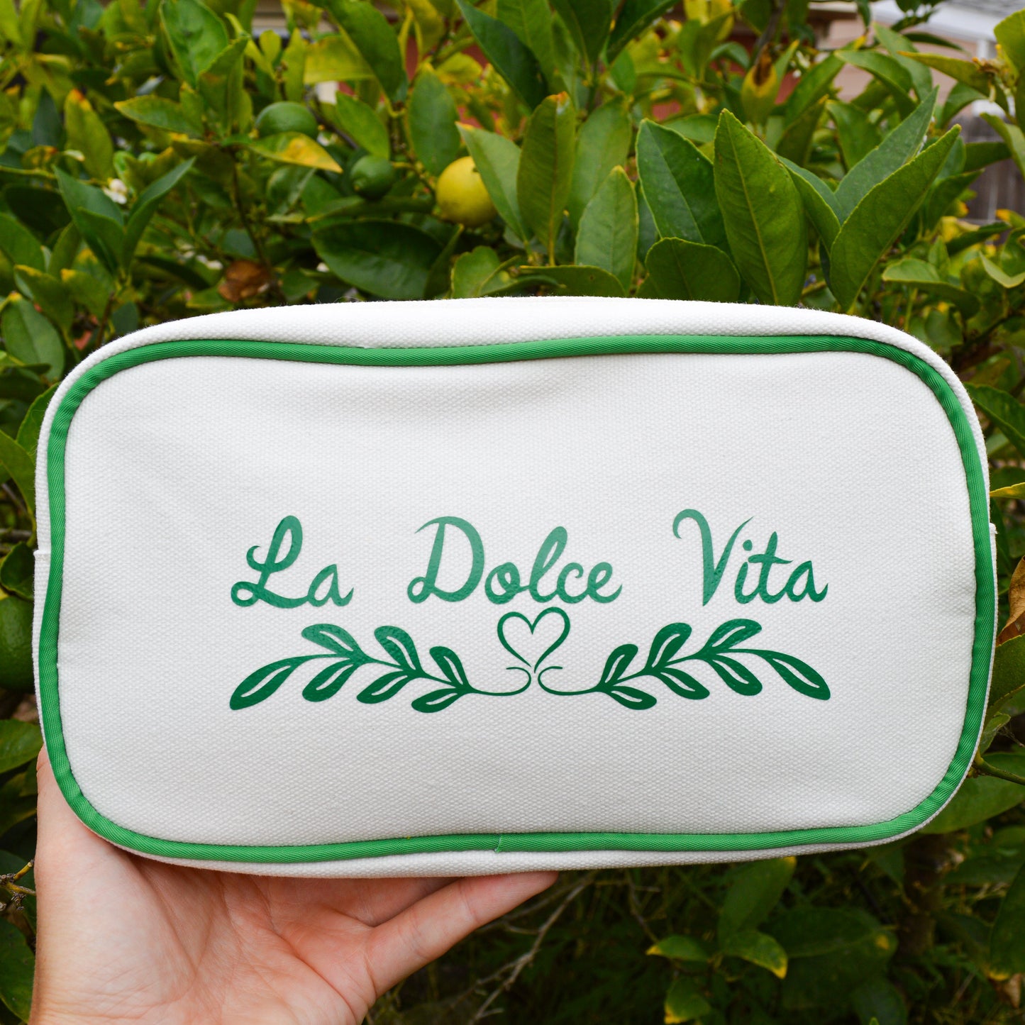 White pouch with 'La Dolce Vita' text held in front of green foliage