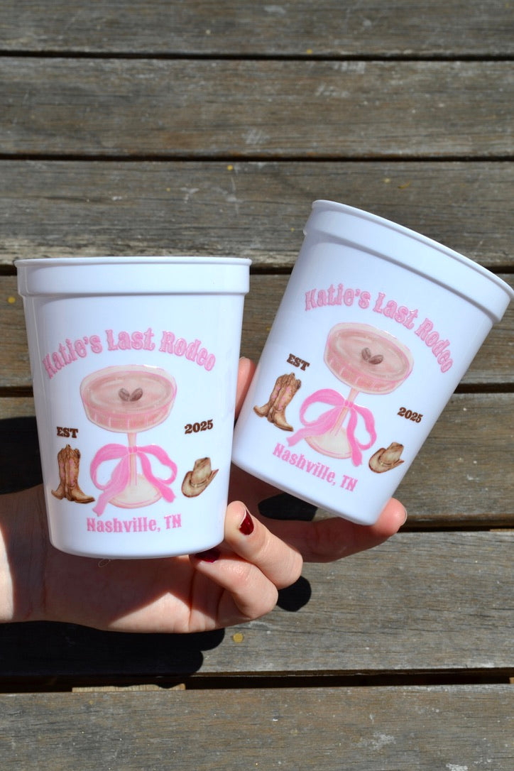 Two white cups with pink designs and text held by a person against a wooden background