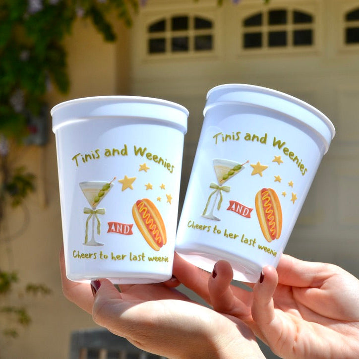 Two white cups with colorful designs and text held by hands against a blurred outdoor background.