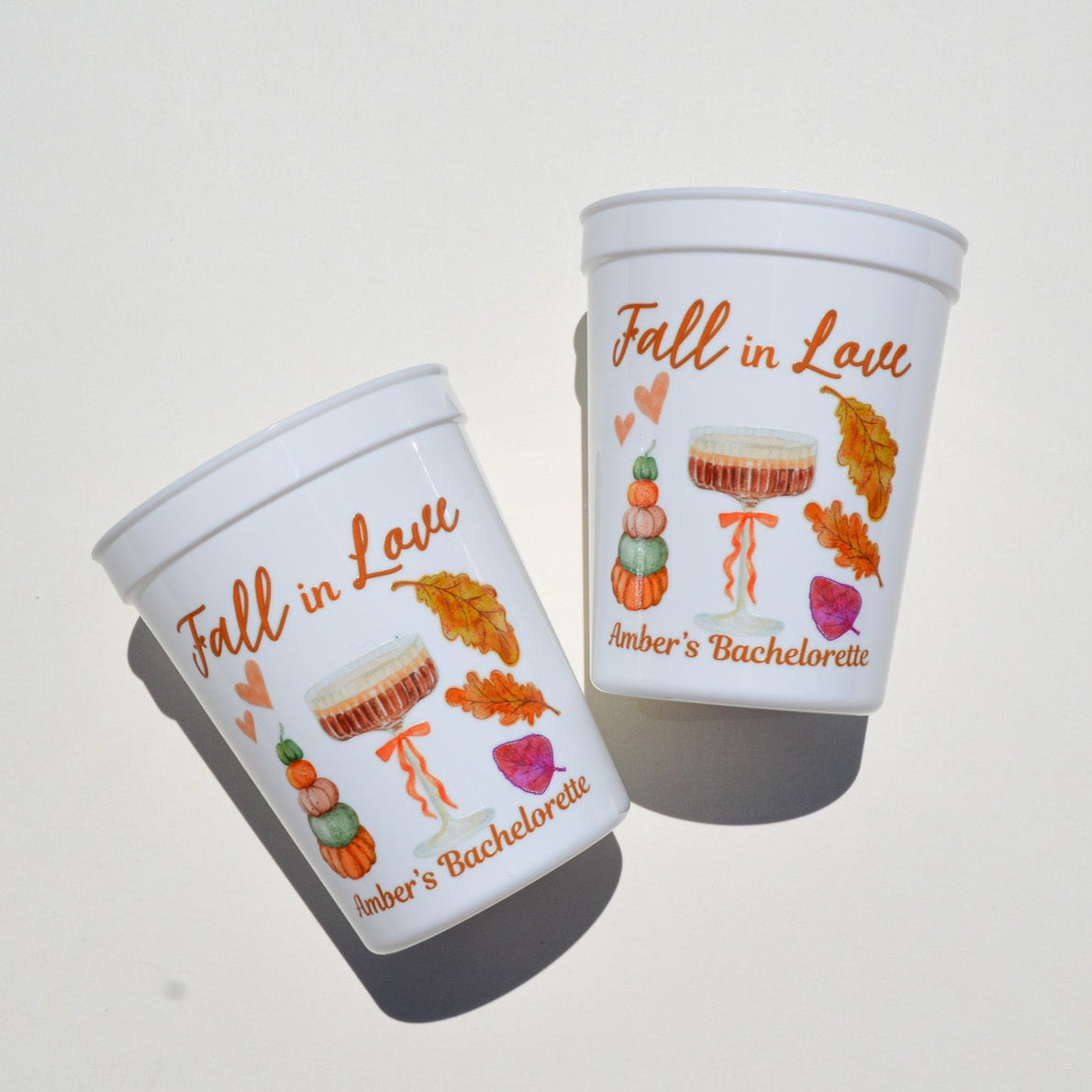 Two white cups with 'Fall in Love' and 'Amber's Bachelorette' text and autumn-themed illustrations on a light gray background.