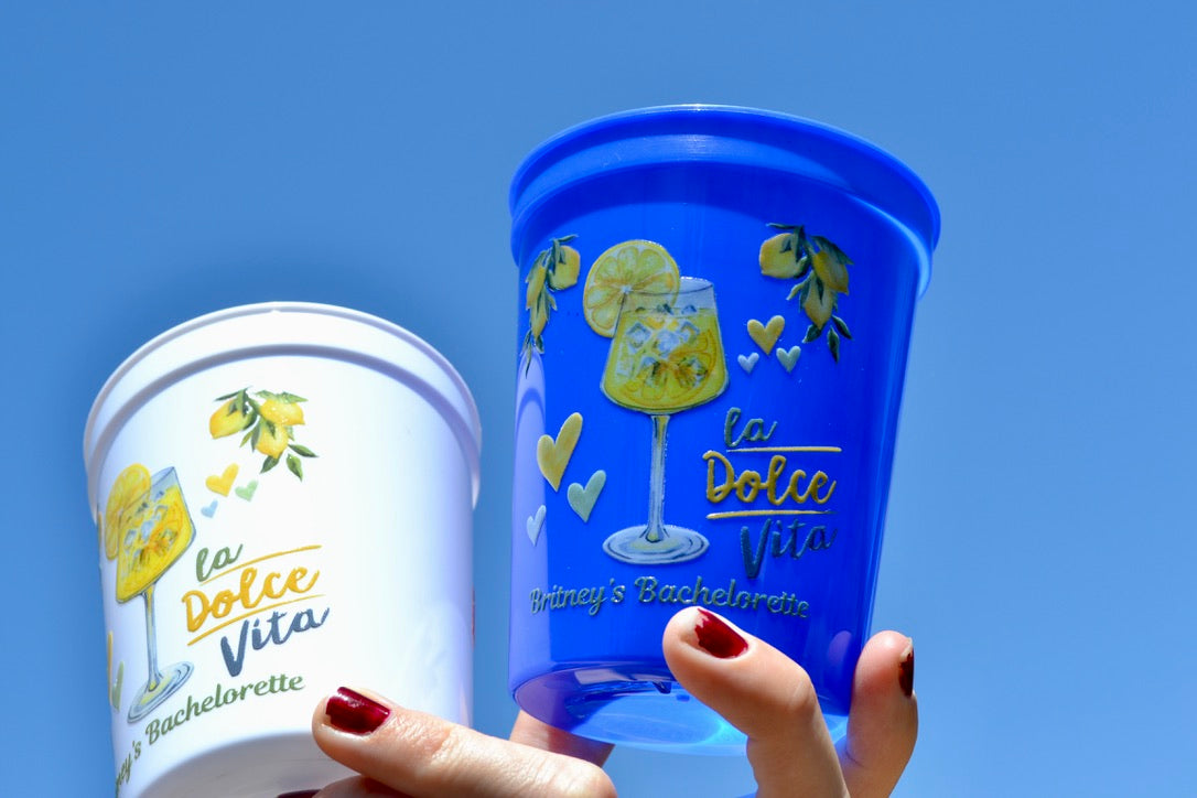 Two cups, one white and one blue, with 'La Dolce Vita' branding against a clear blue sky.