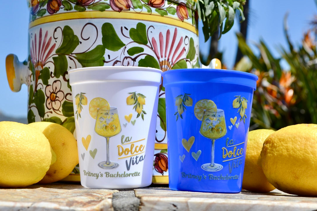 Two decorated cups with lemon designs and text, placed on a wooden surface with lemons and a decorative pot in the background.