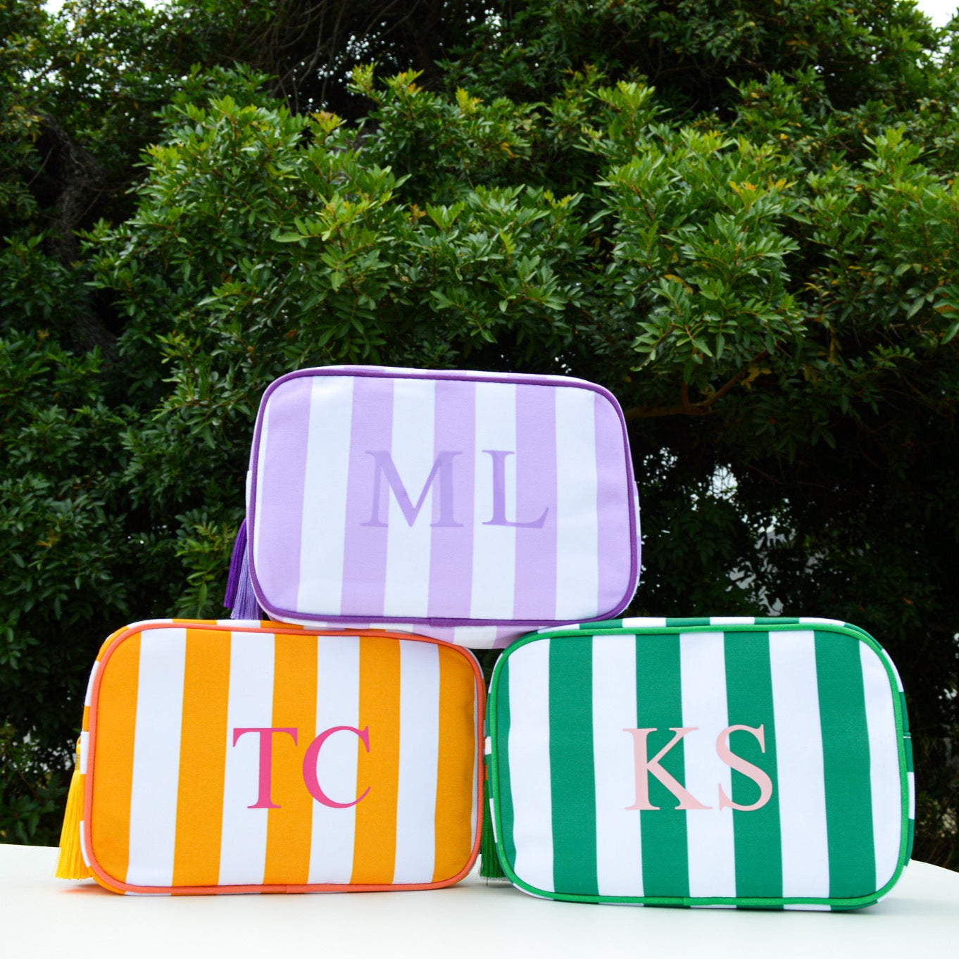 Three striped pouches with monogrammed initials on a white surface with a green leafy background.
