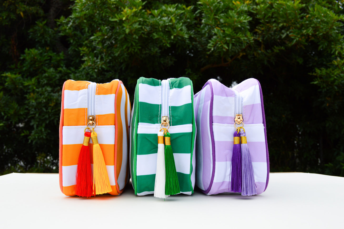 Three colorful zippered pouches with tassels on a white surface with a green outdoor background.