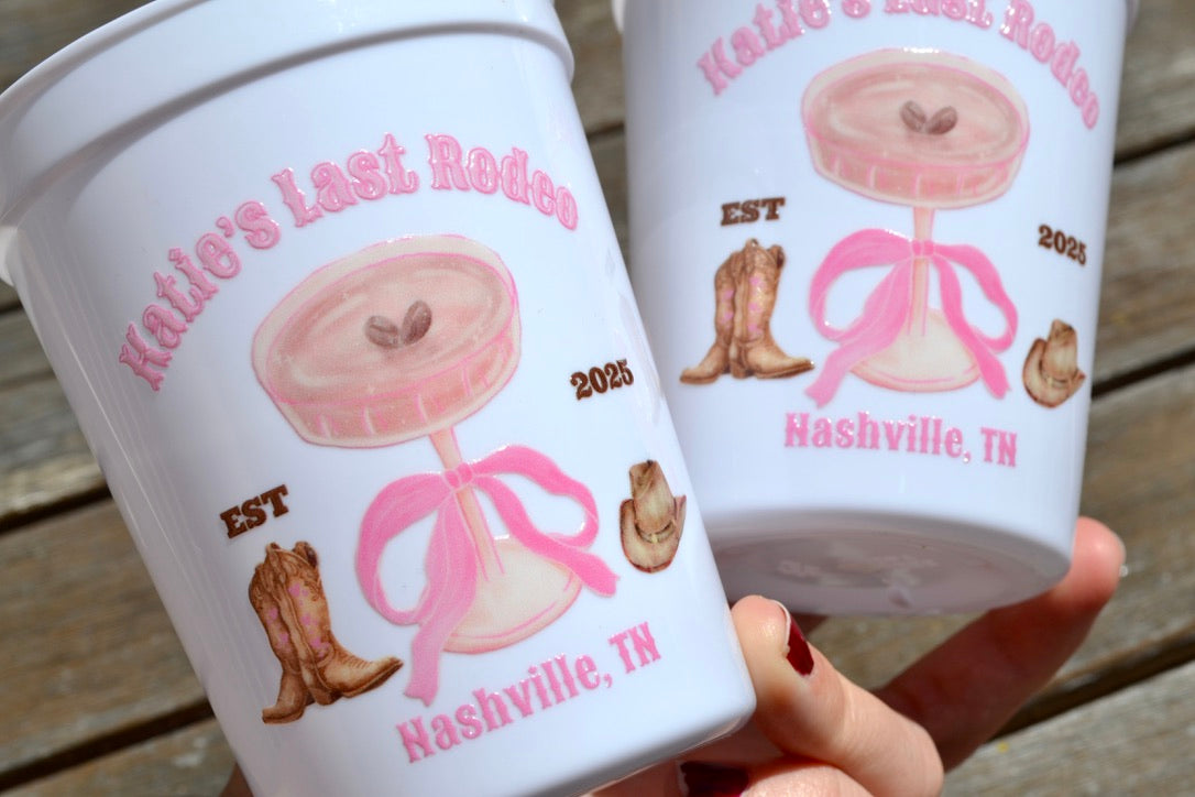 Two white cups with pink and brown graphics and text, held by a hand on a wooden surface.