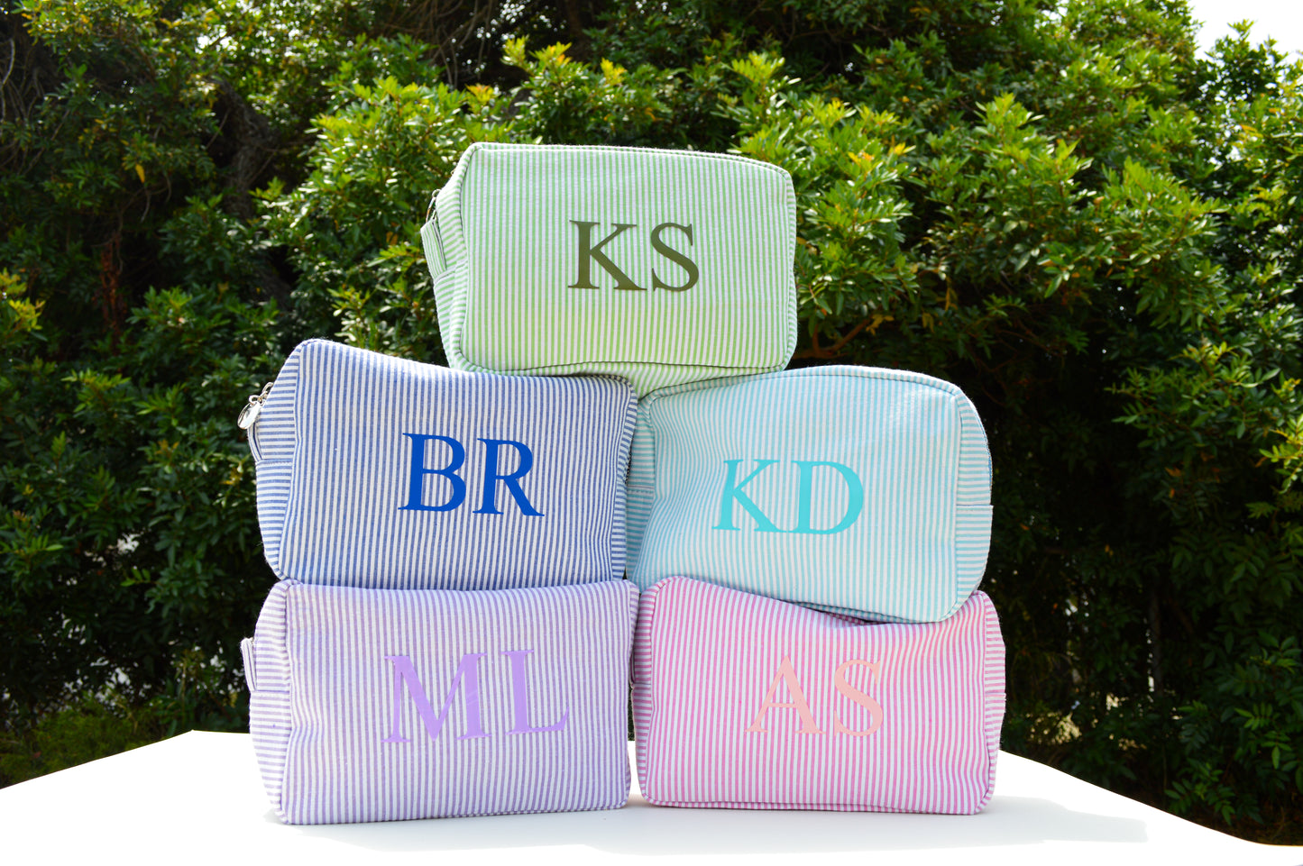 Stack of monogrammed bags with initials 'KS', 'BR', 'KD', 'ML', 'AS' and 'MS' against a green outdoor background.