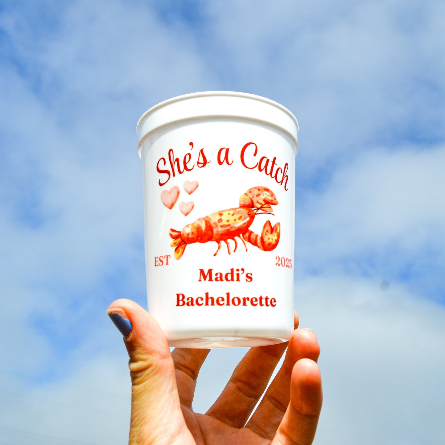 Hand holding a 'She's a Catch' Madi's Bachelorette branded container against a blue sky.