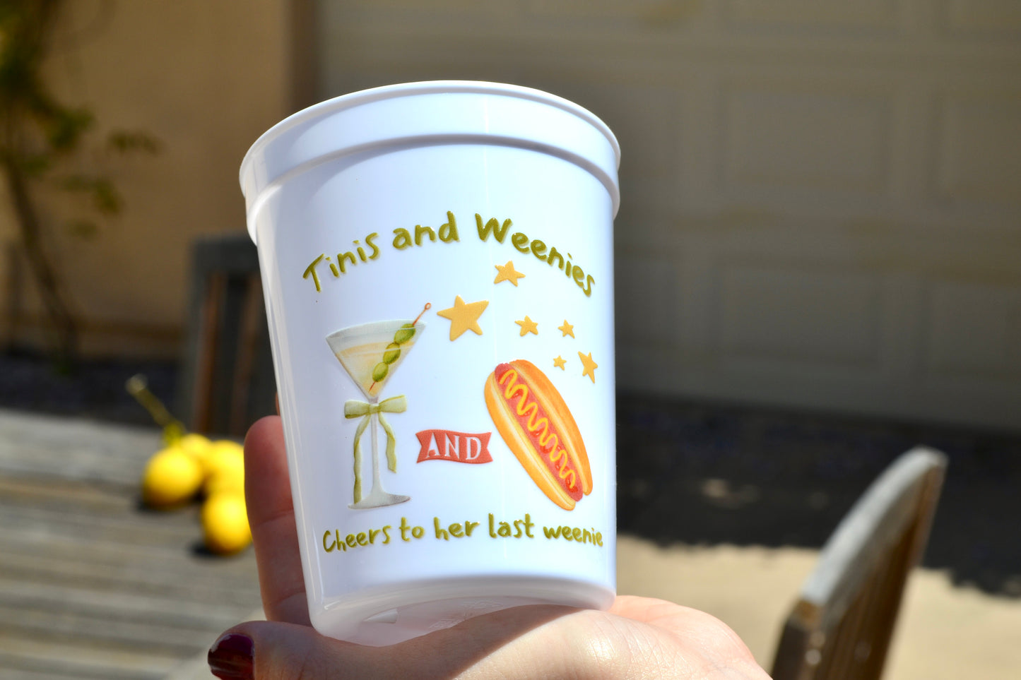 White cup with 'Timis and Weenies' branding and graphics held by a hand outdoors.