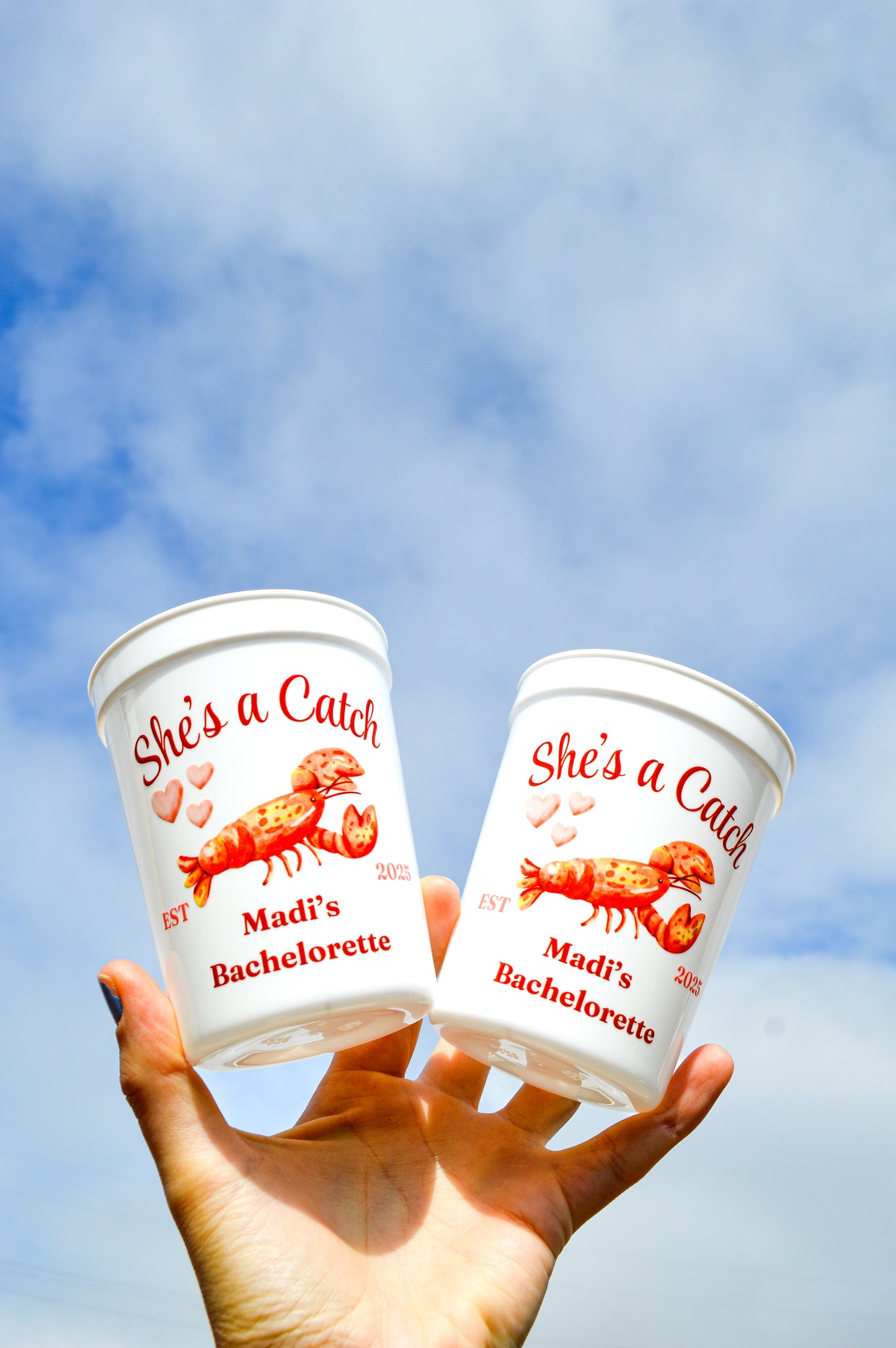 Two 'She's a Catch' Bachelorette-themed cups held against a blue sky.