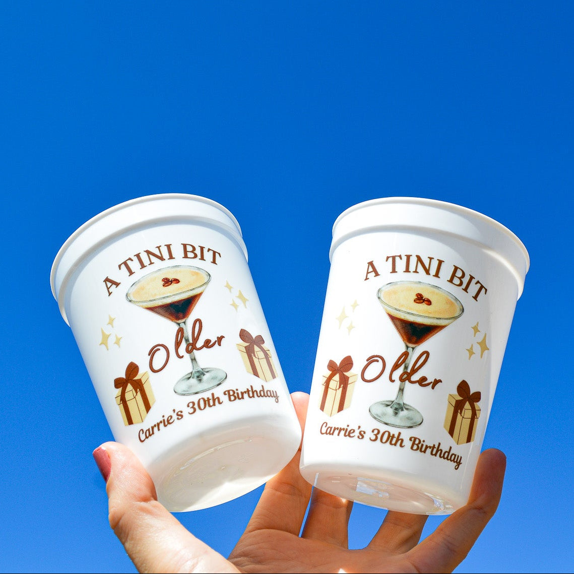 Two white cups labeled 'A Tini Bit' against a clear blue sky.