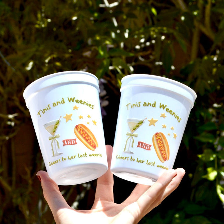 Two plastic cups with 'Tinis and Weenies' text and graphics held by a hand against a blurred green background.