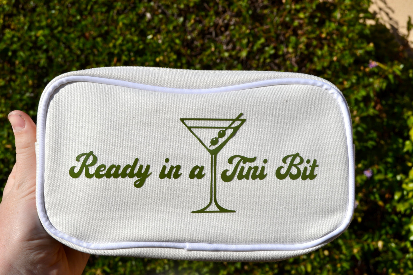 Toiletry bag with 'Ready in a Tini Bit' text and martini glass design held against a green outdoor background.