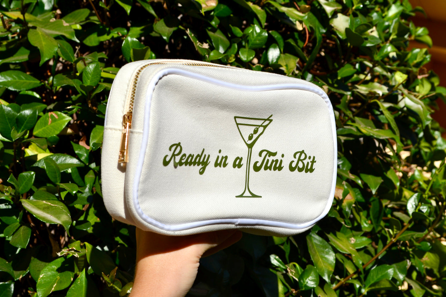 White makeup bag with 'Ready in a Tini Bit' text and martini glass design held in front of green foliage.