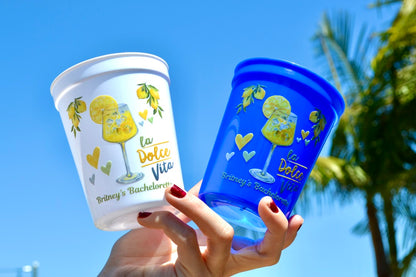 Two branded cups, one white and one blue, held against a clear blue sky with palm trees.