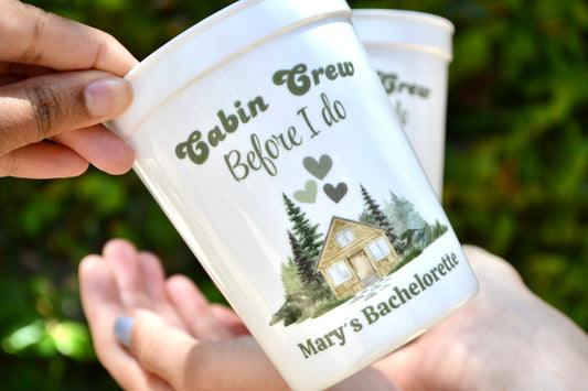Personalized cups with a cabin design and text, held by hands against a blurred green background.