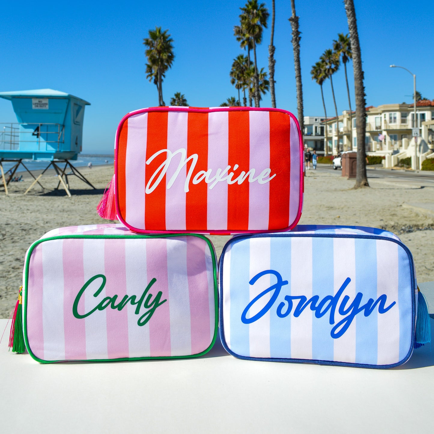 Three striped pouches with names 'Maxine', 'Carly', and 'Jondyn' on a beach.
