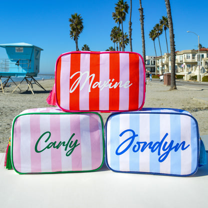 Three striped pouches with names 'Maxine', 'Carly', and 'Jondyn' on a beach.