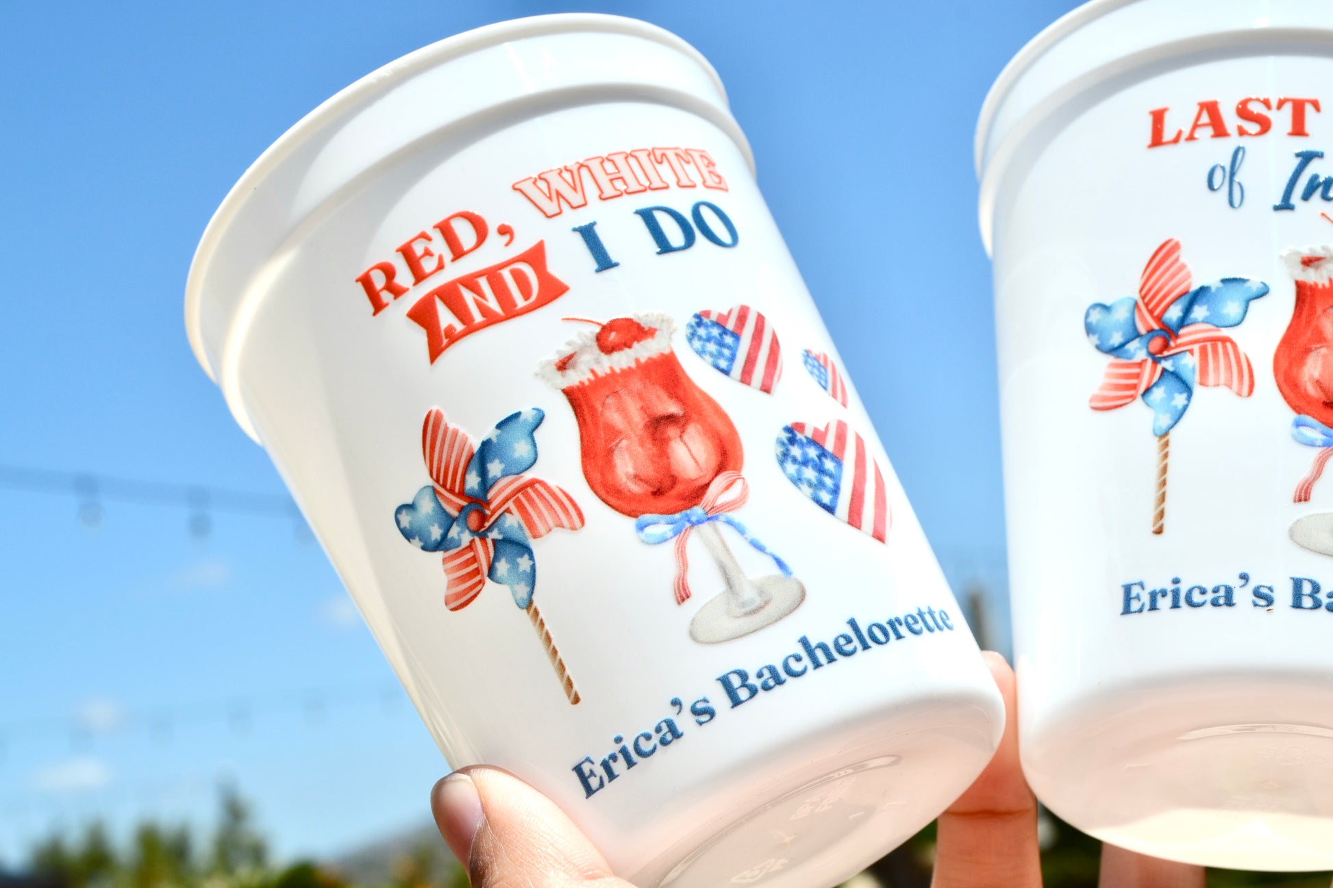Two cups with patriotic designs and text, held against a clear blue sky.