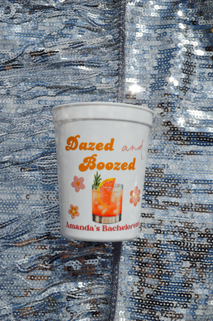 White cup with 'Dazed and Boozed' text and a cocktail graphic on a reflective blue surface.