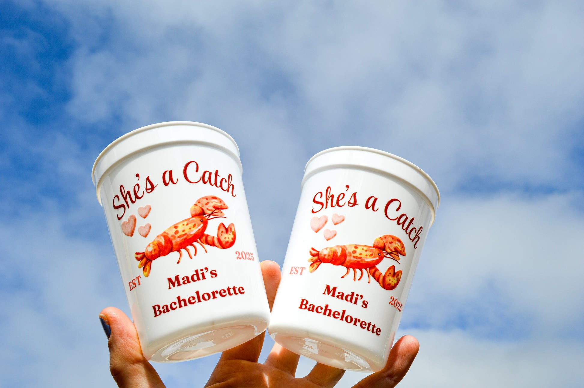 Two paper cups with 'She's a Catch' and 'Madi's Bachelorette' text against a blue sky.