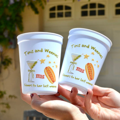 Two white cups with colorful designs and text held by hands against a blurred outdoor background.
