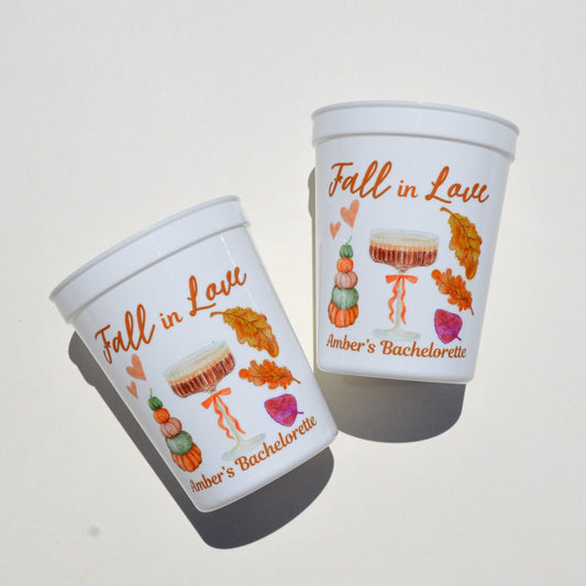 Two white cups with 'Fall in Love' and 'Amber's Bachelorette' text and autumn-themed illustrations on a light gray background.