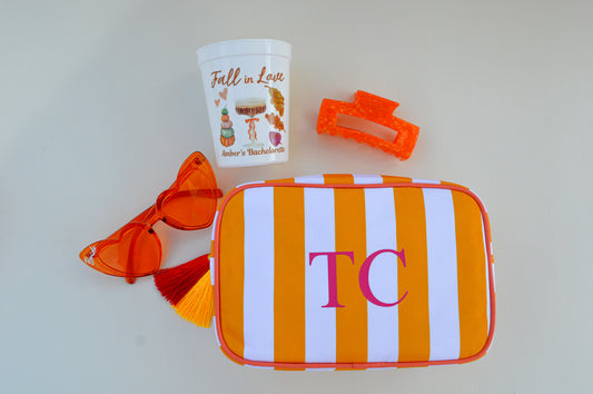Orange and white striped pouch with initials, a small cup, and sunglasses on a light gray background