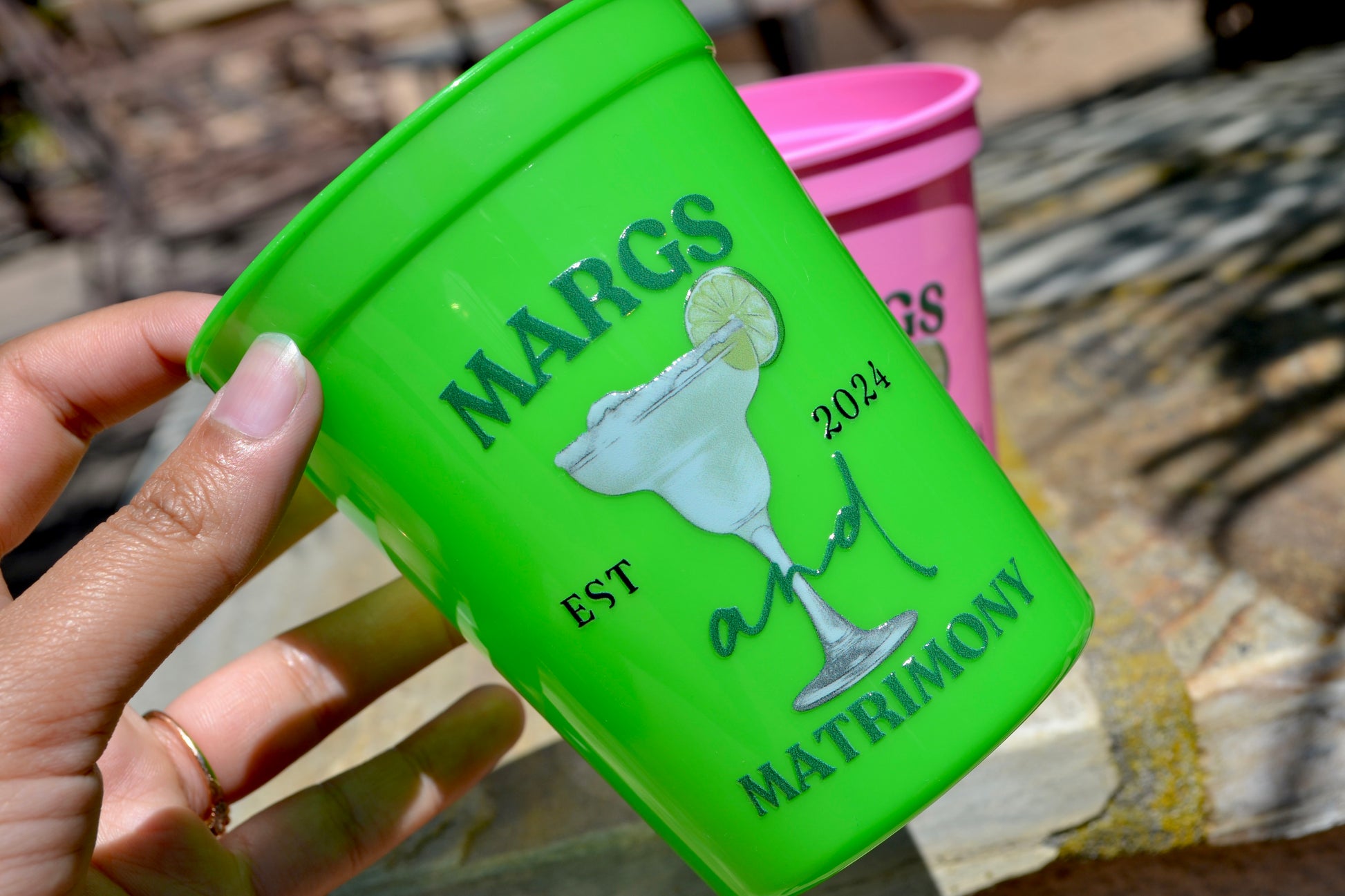Green cup with 'Margs & Matrimony' design held by a hand, with pink cup in the background.