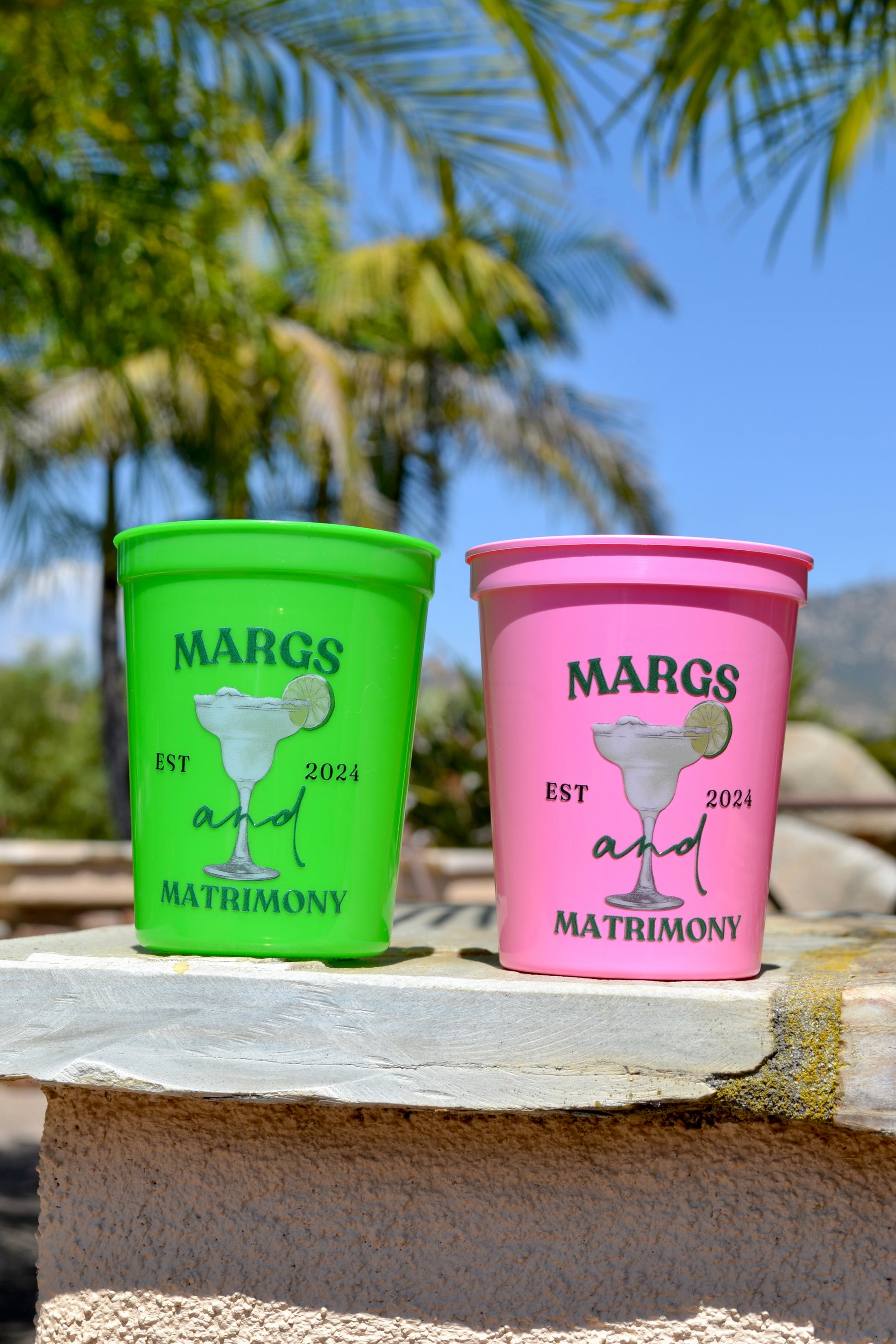 Two colorful cups with 'Margs and Matrimony' text on a stone surface with palm trees in the background.