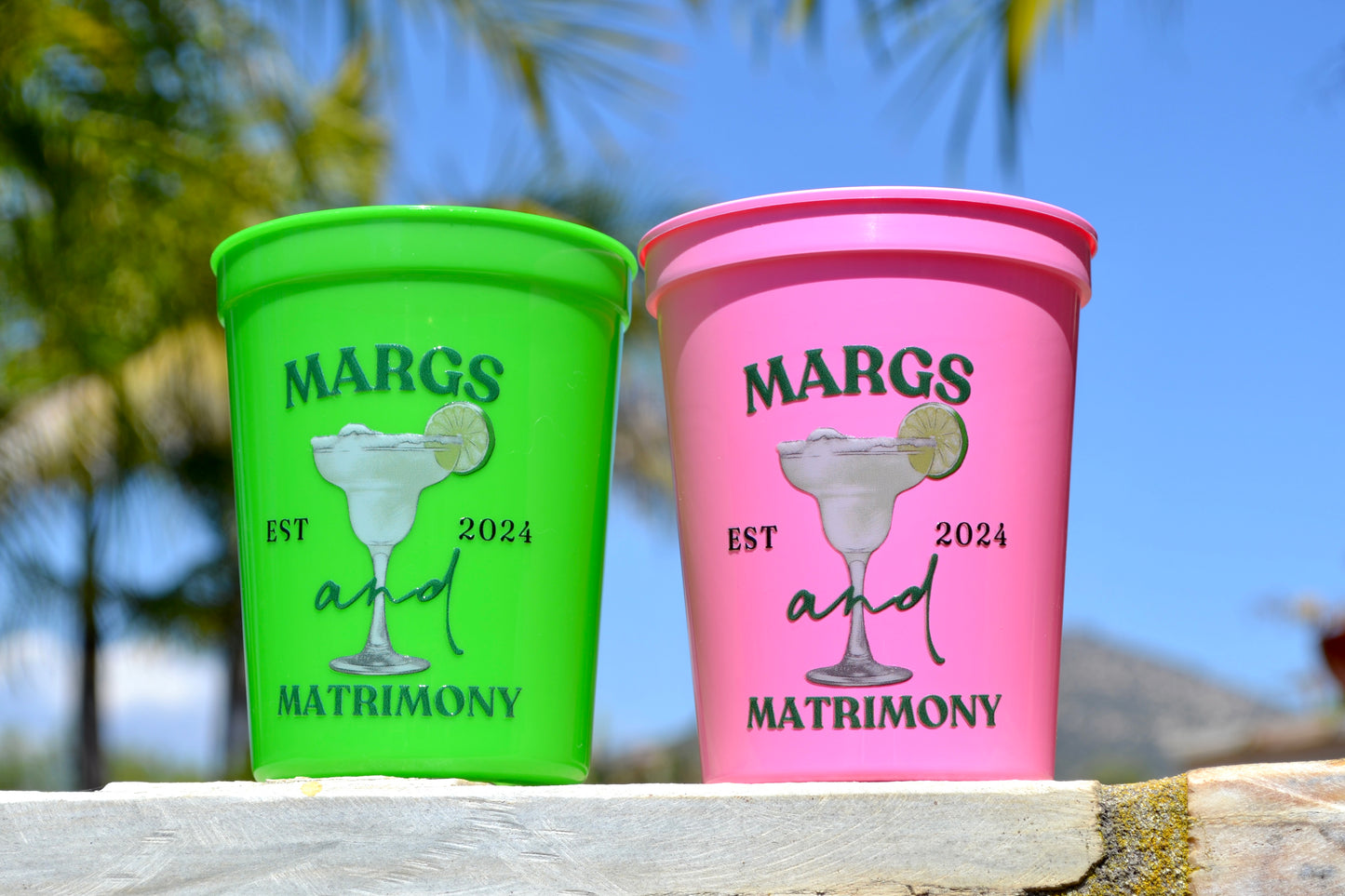 Two colorful cups with 'Margs and Matrimony' text and graphics on a wooden surface with a blurred natural background.