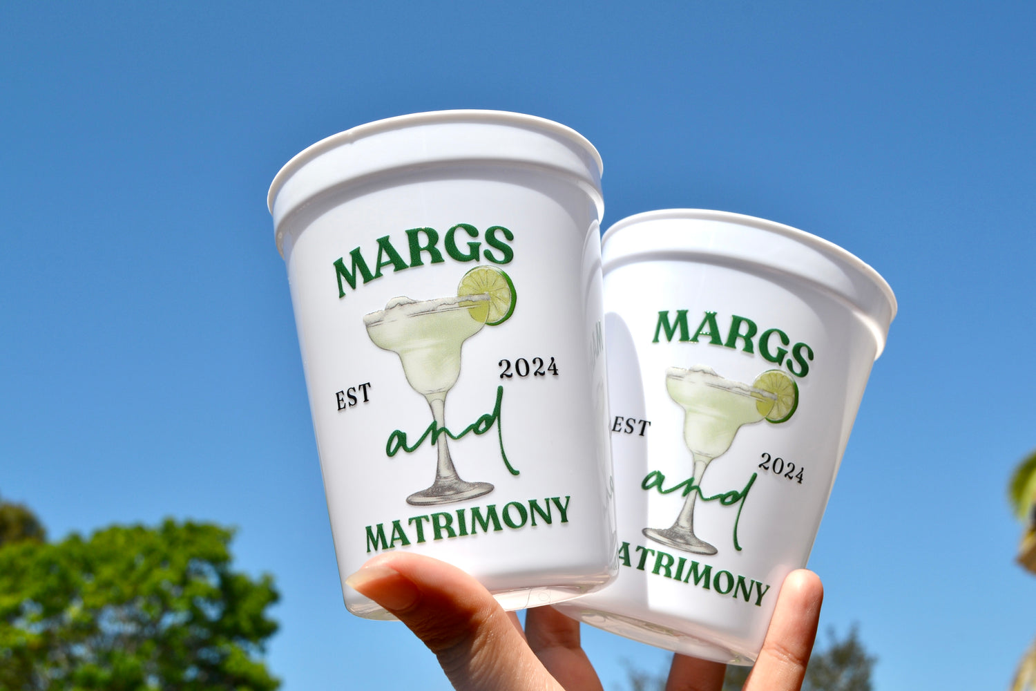Two white plastic cups with 'Margs and Matrimony' design against a clear blue sky.