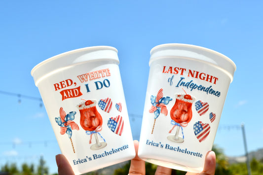 Two plastic cups with festive designs and text, held against a clear blue sky.