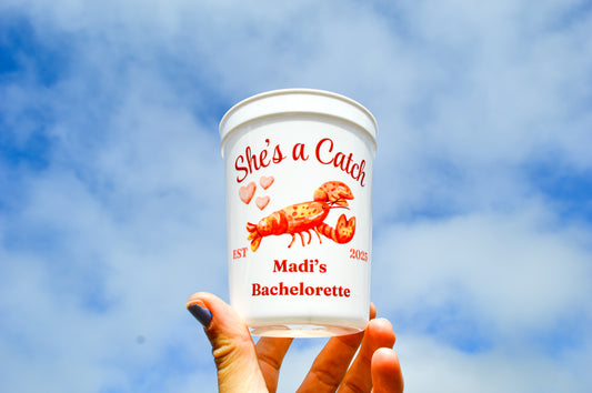 Hand holding a cup with 'She's a Catch' and lobster design against a blue sky.