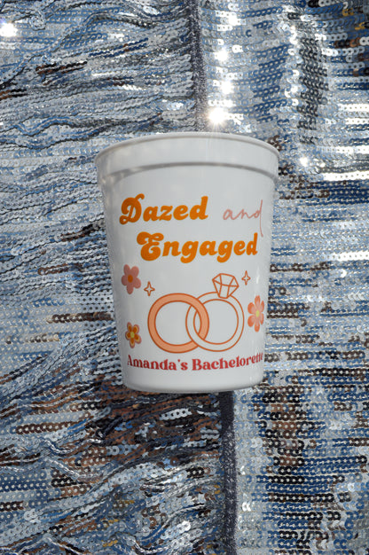 White cup with 'Dazed and Engaged' text and ring design on a reflective surface
