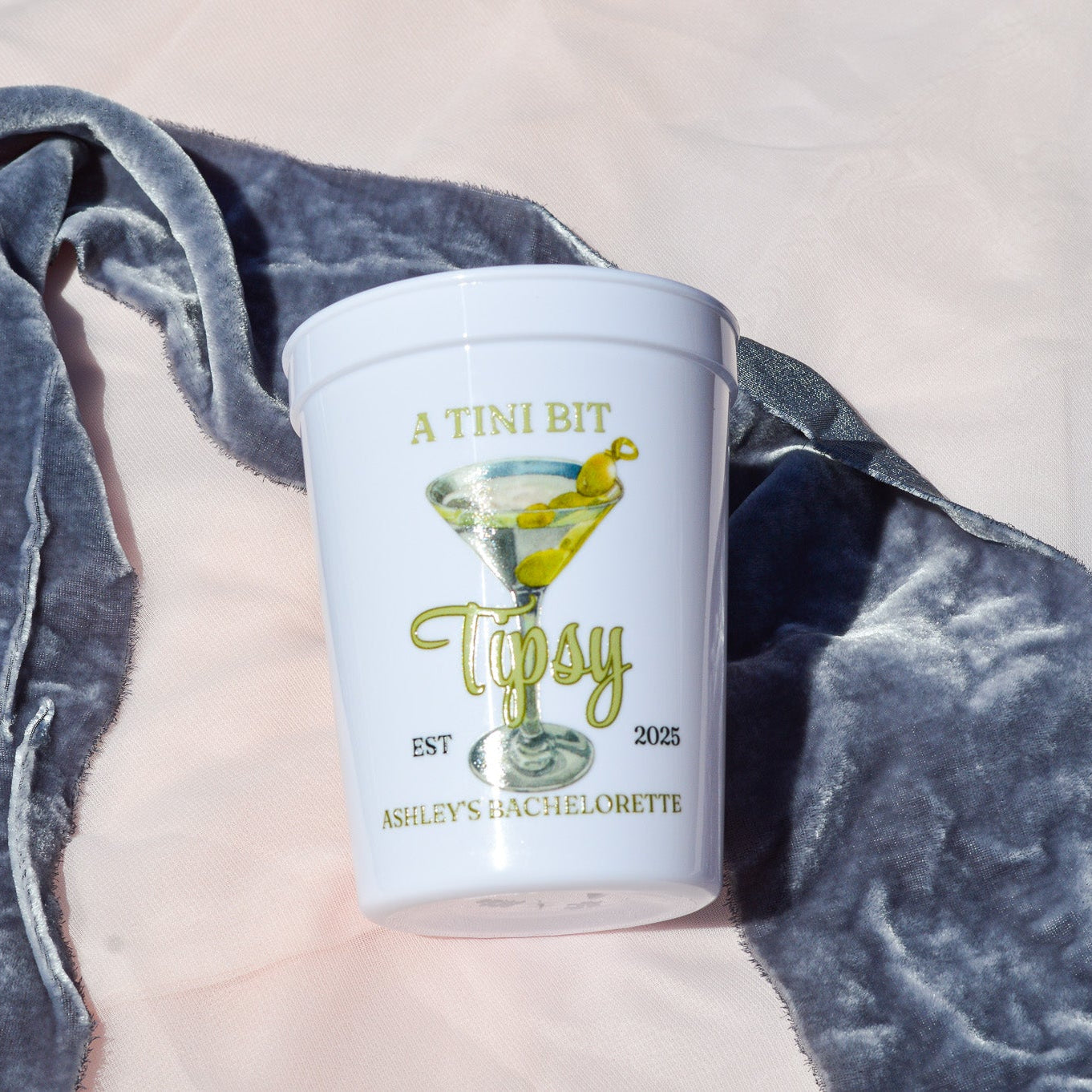 White cup with 'Altiini Bit Tipsy' branding on a gray blanket