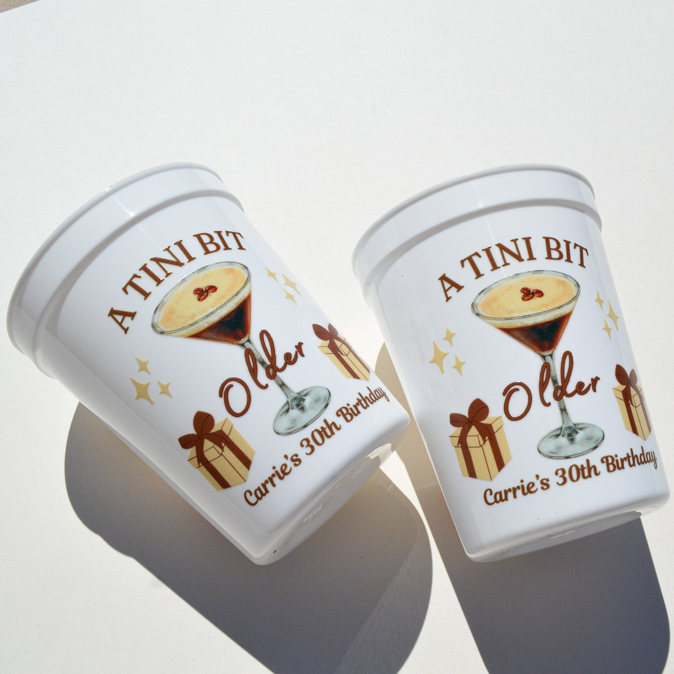 Two personalized white plastic cups A Tini Bit Older with a design and text on a white background