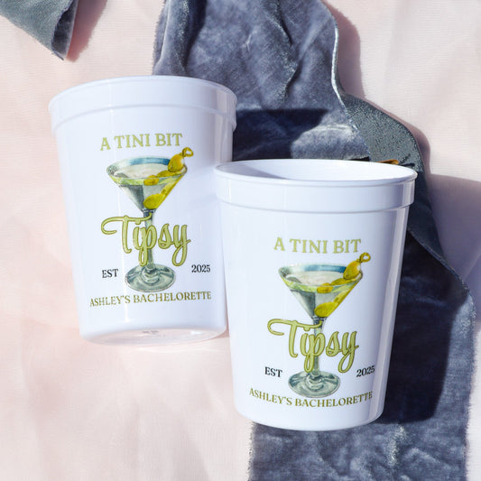 Two white cups with 'A Tini Bit Tipsy' design on a gray fabric background