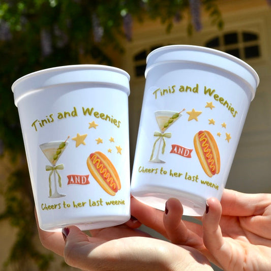 Two white cups with 'Tinis and Weenies' text and graphics held by a person.