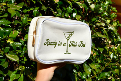 White makeup bag with 'Ready in a Tini Bit' text and martini glass design held in front of green foliage.